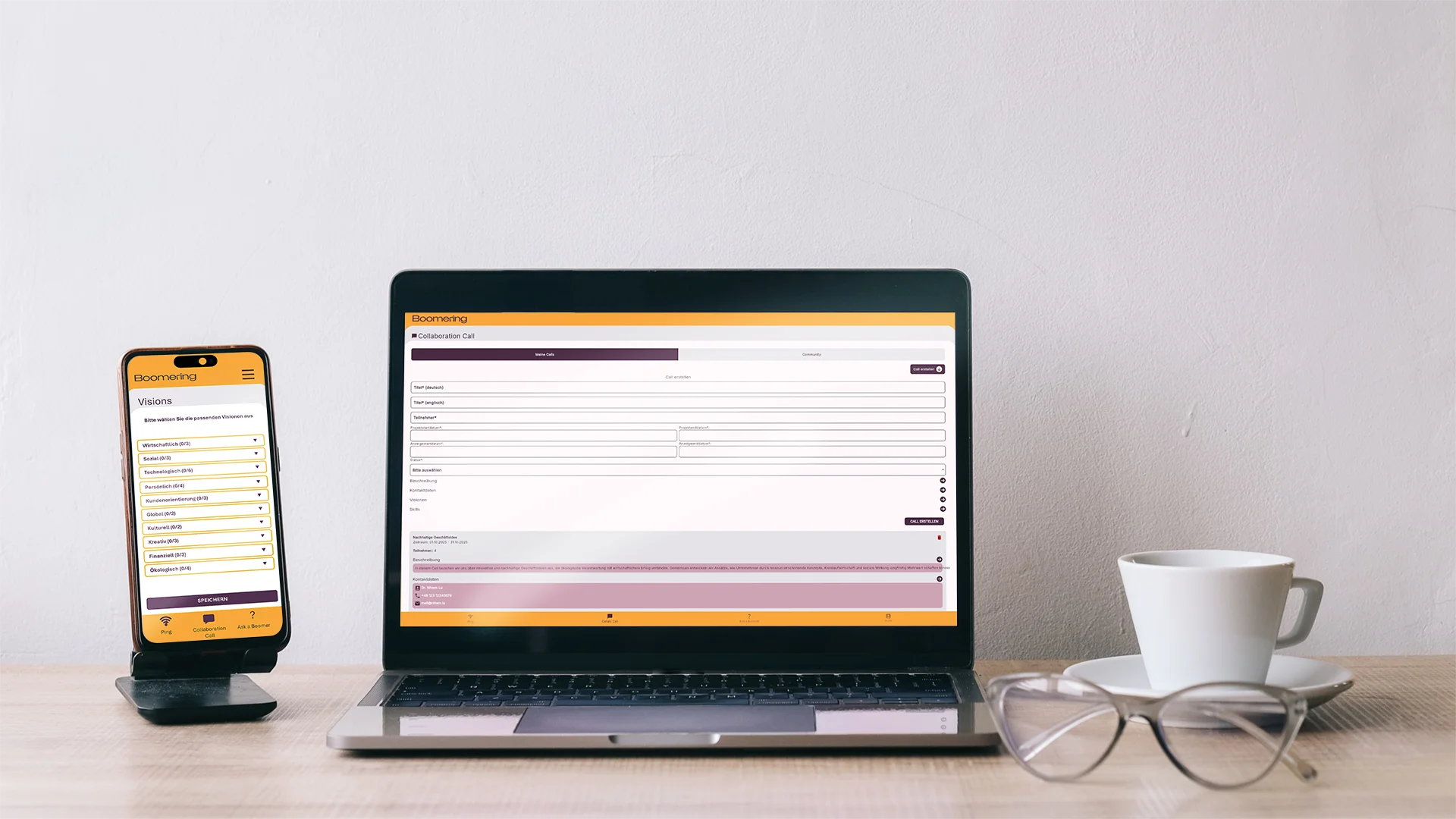 Auf einem Tisch stehen ein Laptop und ein Smartphone mit den Anwendungen aus der App von Boomering, daneben eine weiße Kaffetasse und davor eine graue Brille